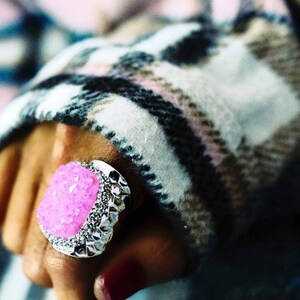 May include: A silver ring with a large, pink, sparkly stone set in the center. The ring is worn on a hand with a plaid sleeve.