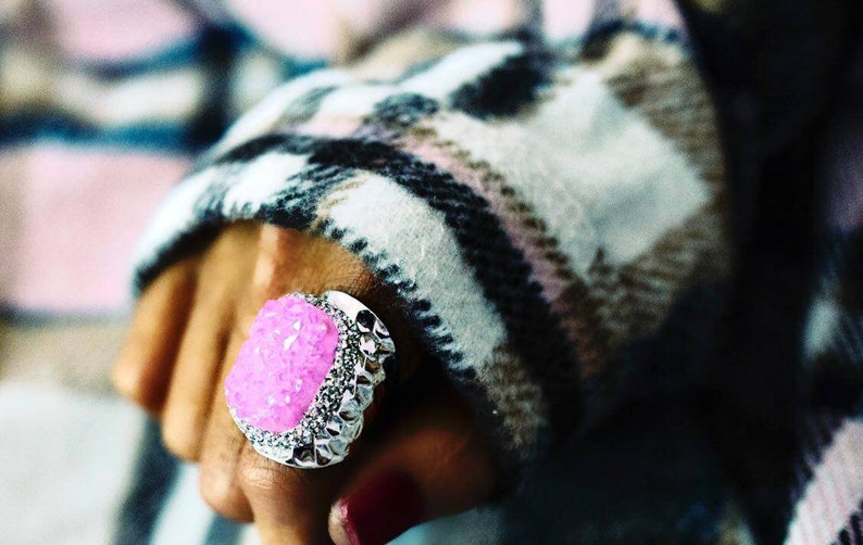 May include: A silver ring with a large, pink, sparkly stone set in the center. The ring is worn on a hand with a plaid sleeve.
