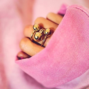 May include: Gold ring with three round balls stacked on top of each other. The ring is worn on a finger.