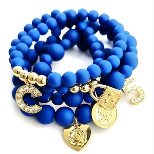 May include: A set of four blue beaded bracelets with gold charms. The charms include a letter "C", a padlock, a heart with a rose design, and a number "5".
