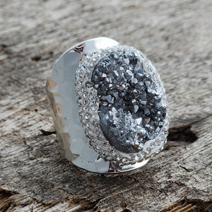 May include: A silver ring with a large, oval-shaped, gray crystal set in a silver bezel. The crystal is surrounded by a row of small, clear rhinestones.