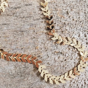 Könnte beinhalten: Eine goldene Kettenkette mit einem Chevron-Muster. Die Kette besteht aus kleinen, miteinander verbundenen Pfeilformen.
