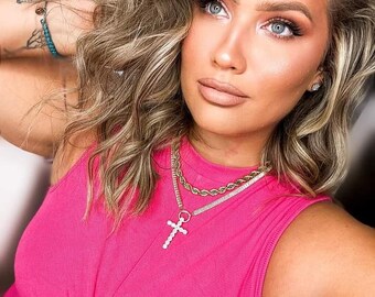 May include: A woman with long blonde hair wearing a bright pink sleeveless top with a knotted front. She is wearing a layered gold chain necklace with a silver cross pendant.