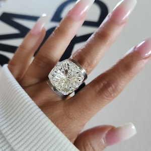 May include: A wide silver band ring featuring a large, clear, faceted cushion-cut stone set in a prominent prong setting. The ring has a polished metallic finish and a substantial, thick band design that sits across the finger.