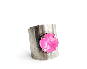 May include: A wide band ring with a distressed, matte silver finish featuring a single, round, faceted hot pink crystal set in a four-prong mount. The metal surface has a textured, weathered appearance, contrasting with the bright, reflective pink stone.