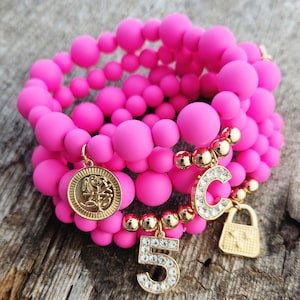 May include: A set of four pink silicone bracelets with gold charms. The charms include a rose, the letter C, a padlock, and the number 5. Each charm is decorated with rhinestones.