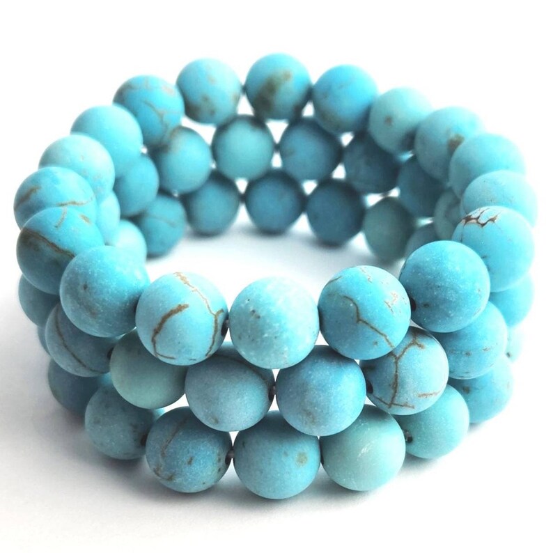 Puede incluir: Una pulsera de tres hileras compuesta por cuentas redondas de acabado mate en color turquesa. Cada cuenta presenta un patr&oacute;n veteado de color marr&oacute;n oscuro en su superficie, lo que crea una apariencia texturizada en toda la pieza circular.