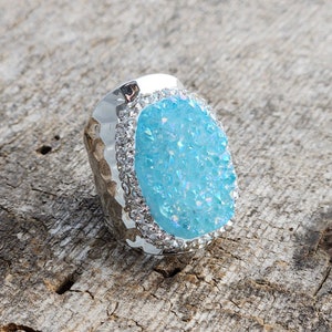 May include: A silver ring with a large, oval-shaped blue gemstone surrounded by smaller clear rhinestones.