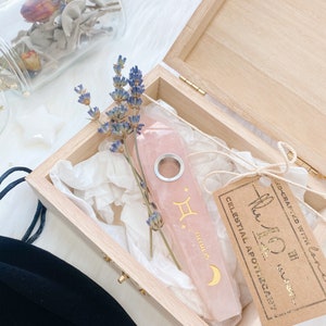 May include: A pink crystal pipe with a gold Gemini zodiac symbol and crescent moon design. The pipe is in a wooden box with a brown paper tag that reads "Celestial Apothecary".