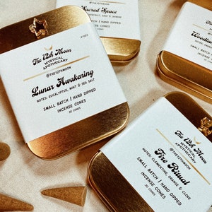 以下が含まれることがあります： 白いラベルが付いた金色の香炉が4つ。ラベルには「The 12th Moon Mystical Apothecary」というテキストと、香りの名前「Sacred Space」、「Lunar Awakening」、「Fire Ritual」、「Woodland」が書かれています。各香炉には30個の香炉が含まれています。