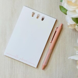 Könnte beinhalten: Ein weißer Notizblock mit einem rosa handschriftlichen Namen "nikki gaharan" am unteren Rand. Oben auf dem Notizblock befinden sich drei Illustrationen von Kaffeegetränken. Ein rosa Stift liegt neben dem Notizblock.