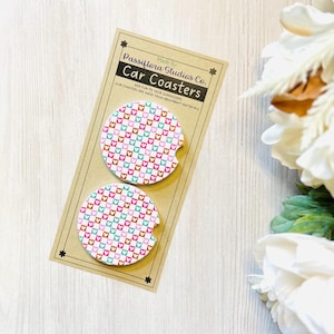 May include: Two car coasters with a colorful heart pattern, packaged on a brown card. The coasters are white with a repeating pattern of pink, teal, and brown hearts. The card reads "Passiflora Studios Co. Car Coasters."