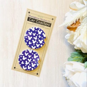 May include: Two car coasters with a dark blue background and a white Mickey Mouse pattern. The coasters are displayed on a cardboard backing with the text "Car Coasters" and "Passiflora Studios Co."