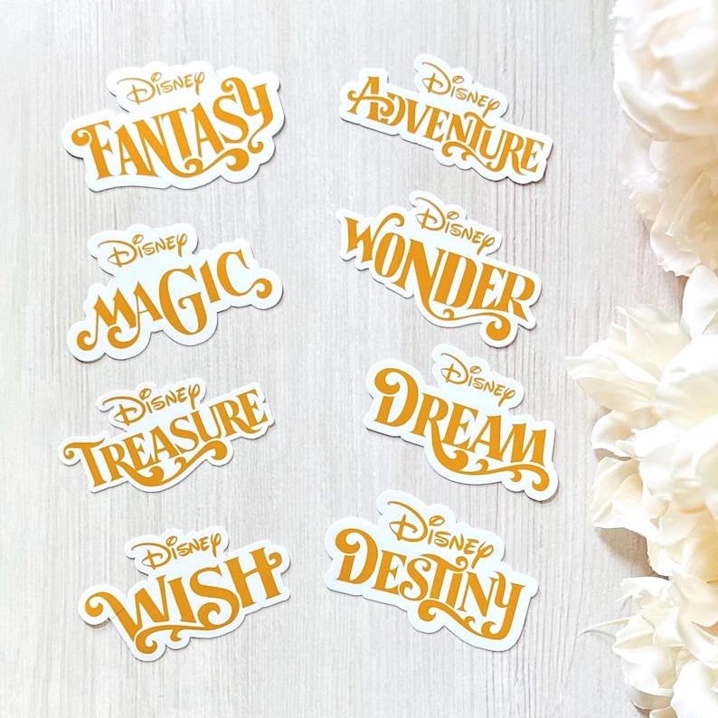 Pu&ograve; includere: Una collezione di adesivi Disney con parole come "Fantasy", "Adventure" e "Magic" in un font arancione. Ogni adesivo ha un bordo bianco ed &egrave; disposto su una superficie con venature di legno chiaro.