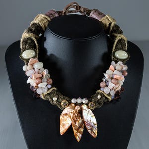 Op de afbeelding: Een handgemaakte ketting met een gevlochten bruin koord, versierd met een verscheidenheid aan stenen in roze, witte en bruine tinten. De ketting bevat drie iriserende, bladvormige hangers en wordt gepresenteerd op een zwarte sieradenstandaard.