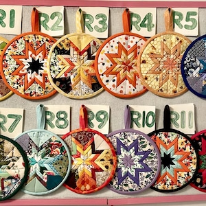 May include: A collection of twelve colorful, round quilted pot holders with starburst designs. Each pot holder features a different fabric pattern and color scheme, with a loop for hanging. The pot holders are displayed on a pink-framed board.
