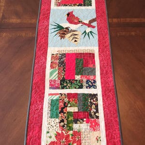 May include: A rectangular quilted table runner featuring a cardinal bird design. The runner has a red background with a gray border and white accents. The design includes patchwork squares with floral and geometric patterns.