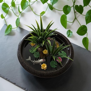 May include: A round, dark brown ceramic bowl filled with water, containing two green spiky plants, several small yellow and pink floating flowers, green lily pads, and a small, detailed white and grey koi fish figurine swimming inside.