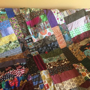 May include: A patchwork quilt made from a variety of colorful fabrics, featuring a mix of floral, geometric, and abstract patterns. The quilt is sewn together with a simple grid pattern and has buttons sewn on each square.