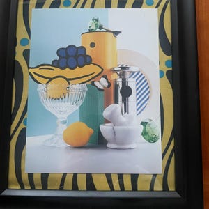 May include: Framed art print featuring a still life composition. The artwork includes a yellow teapot, a glass bowl with a lemon, a marble mortar and pestle, and a bunch of grapes. The background has a blue and white color scheme.