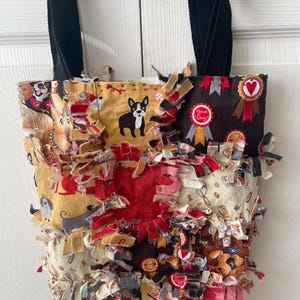 May include: A handmade tote bag with a patchwork design featuring various dog breeds and decorative ribbons. The bag has a black strap and is embellished with frayed fabric strips in red, yellow, and brown.