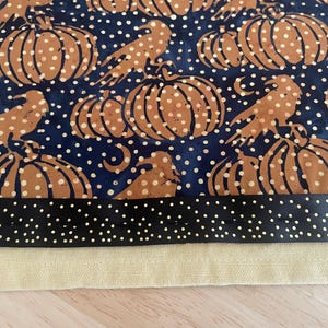 May include: A fabric with a repeating pattern of brown pumpkins and birds on a navy blue background with white polka dots. A black border with white polka dots and a tan fabric edge are at the bottom.