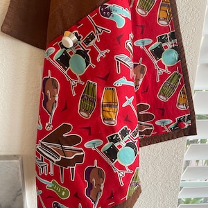 May include: A red fabric kitchen towel with a brown border and a pocket. The pocket is lined with a red fabric with a pattern of musical instruments, including a piano, drums, guitars, and a cello.