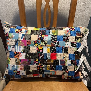 May include: A patchwork pillow with a variety of colorful fabrics, including blue, red, green, and black. The pillow has a square pattern and is tied with fabric ties.