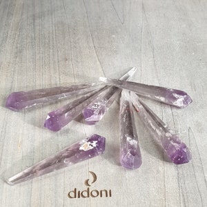 May include: A group of seven amethyst crystal points, each with a pointed tip and a translucent purple hue. The crystals are arranged in a fan shape on a light wood surface.