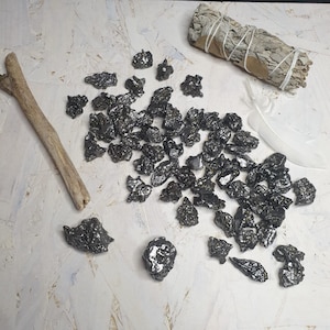Puede incluir: Una colección de piedras de tectita negras, también conocidas como moldavite, sobre una superficie blanca. Las piedras son de varias formas y tamaños, y algunas tienen un acabado brillante y metálico.