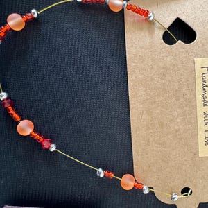 May include: A delicate beaded bracelet featuring orange and red beads interspersed with silver-colored beads. The bracelet is strung on a thin, gold-colored wire. A brown tag with the words "Handmade with Love" is attached.