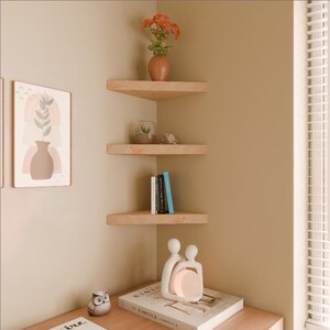 May include: Three wooden corner shelves with a light wood finish. The top shelf has a small orange flower arrangement in a terracotta pot. The middle shelf has a small glass terrarium with a succulent plant. The bottom shelf has a stack of books and a white ceramic sculpture of two people.