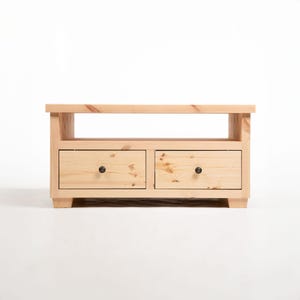 May include: A light-coloured wooden TV stand with two drawers and a shelf. The drawers have black knobs. The stand is made of unfinished wood and has a simple, modern design.