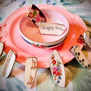 May include: A collection of eight vintage porcelain hair clips with floral and bunny designs. The clips are arranged on a pink plate with a small bowl that says "Happy Times".