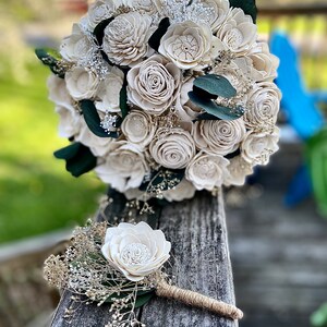 May include: A white wooden flower bouquet with greenery and a matching boutonniere. The bouquet is made of sola wood flowers and is perfect for a rustic wedding.