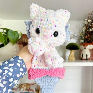 May include: A handmade crochet mermaid cat doll. The doll features a white head and body with colorful speckles, black eyes, and pink accents. It has a pink bow and a light blue mermaid tail. The doll is approximately 12 inches tall.