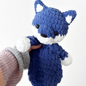 May include: A handmade blue and white crochet fox toy. The fox has a textured, plush appearance with white accents on the ears, paws, and snout. It features black safety eyes and a small black nose. The fox is being held.