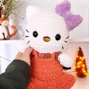 May include: A white crocheted Hello Kitty plush toy wearing an orange dress with a purple bow. The toy is being held by a hand.