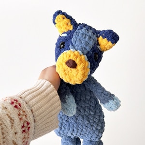May include: A handmade blue and yellow crochet stuffed animal. The plush toy has a light blue body, darker blue patches, and yellow accents on the ears and snout. The toy is held against a white background.