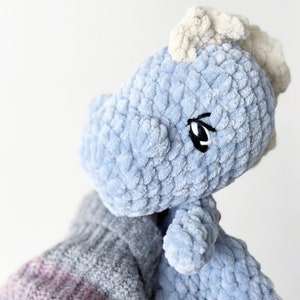 May include: A light blue crocheted plush toy with a textured, soft yarn finish. The creature features a cream-colored mane along the top of its head and embroidered black and white eyes. The plush has a rounded snout and a small, soft body.