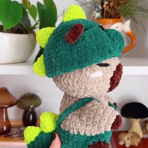 May include: A handmade crochet dinosaur plushie in shades of green, brown, and tan. The plushie features a dinosaur hood with lime green spikes and brown accents. The toy is held in a person's hand.