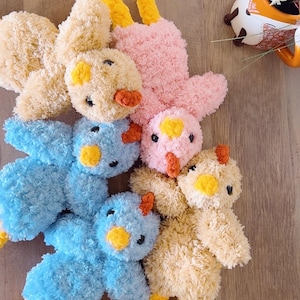 May include: A collection of handmade plush chicks in various colors, including pink, blue, and tan. Each chick features yellow feet, a yellow beak, and small black eyes. The soft toys are arranged on a wooden surface.