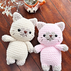 May include: Two handmade crochet cat plushies. One is cream-colored with gray accents, the other is pink with gray accents. Both have black eyes, brown noses, and small whiskers. They are positioned on a wooden surface.