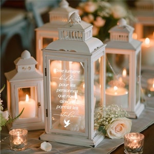 May include: White lantern with a glass panel featuring the text "Cette lumière brille pour les personnes que nous aimons parmi les étoiles" and a white dove graphic. The lantern is decorated with a white candle inside.