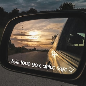 Può includere: Specchietto retrovisore laterale di un'auto che riflette un tramonto su una strada. Il testo "we love you, drive safe" è scritto sullo specchietto. Il cielo è un mix di arancione, giallo e blu.