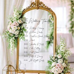 Op de afbeelding: Een goudomrand spiegel met een bruilofttijdlijn, versierd met bloemstukken van witte en roze rozen. De spiegel staat op een gouden standaard, met brandende kaarsen in lantaarns aan de basis. De tekst luidt "The Wedding Timeline".