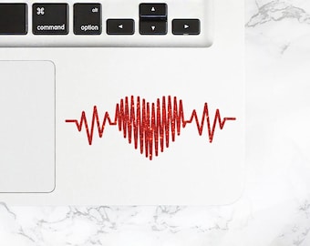 Könnte beinhalten: Ein weißer Laptop mit einer roten herzförmigen EKG-Grafik auf dem Trackpad. Der Laptop befindet sich auf einer weißen Marmorfläche mit einer rosa Tasse und einer Perlenkette.