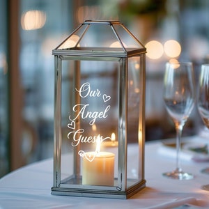 May include: A clear glass lantern with a silver metal frame and a candle inside. The lantern has a white vinyl decal that reads "Our Angel Guests" with hearts around the text.