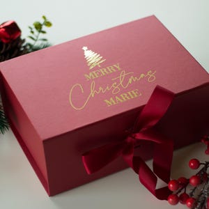 Puede incluir: Una caja de regalo roja con un árbol de Navidad dorado y las palabras "MERRY Christmas MARIE" impresas en la tapa. Una cinta roja está atada alrededor de la caja. Piñas y bayas decorativas rodean la caja.