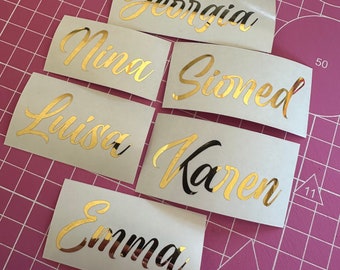 May include: Six gold foil name stickers on white paper. The names are: Georgia, Nina, Sianed, Luisa, Karen, and Emma.